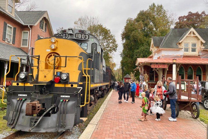 Vintage train on tracks with people dressed in costumes on platform near brick building.