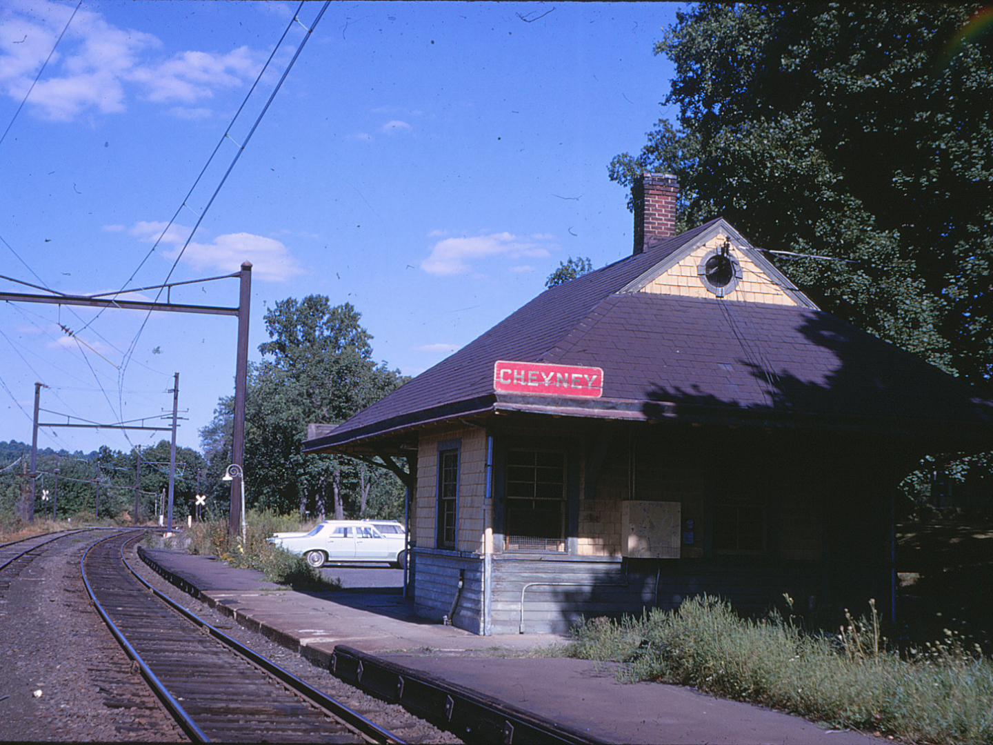 Rural train station with sign, tracks, and parked car on a sunny day.