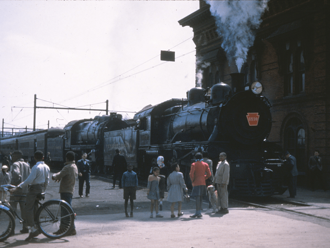 Steam locomotives at a station with people and bicycles nearby.