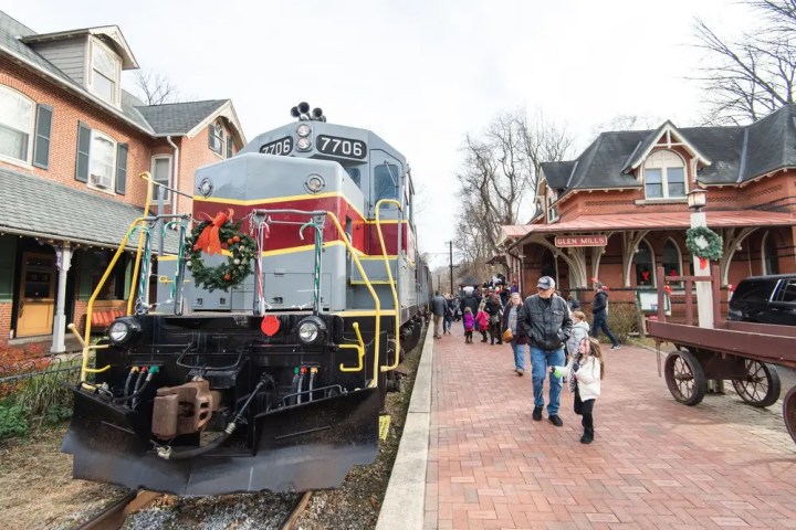 Train with wreath at station, people walking on platform, brick buildings nearby.